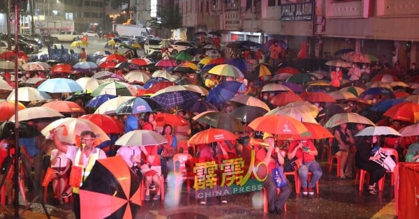 台下出席者举起雨伞或穿上雨衣，继续留在现场以表支持。